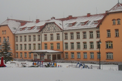 W oddali widać plac zabaw, w tle budynek główny szpitala, wszystko pokryte jest śniegiem.