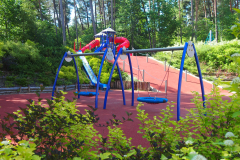 Zdjęcie przedstawia plac zabaw który mieści się na terenie szpitala. Na pierwszym planie widać dwie bujawki w niebieskim kolorze, w tle widać czerwoną zjeżdżalnie. Teren otacza las.