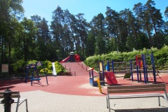 Po prawej stronie czerwono-niebieski plac zabaw ze ścianką wspinaczkową, piaskownicą oraz zjeżdżalnią. Po lewej stronie dwie bujawki, w tle las.
