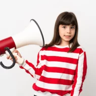 zdjęcie przedstawia dziewczynkę w ciemno brązowych włosach która ubrana jest w białą bluzkę z czerwonymi pasami, w ręku trzyma megafon