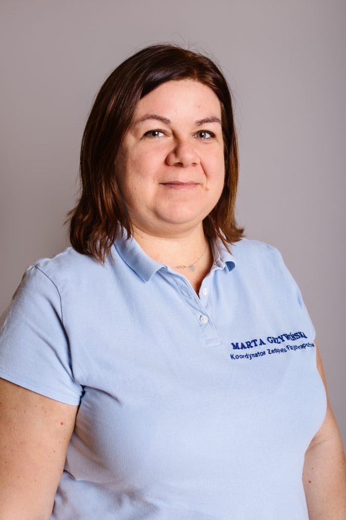 mgr Marta Grzywińska Koordynator zespołu fizjoterapeutów. Kobieta w brązowych włosach, w niebieskiej koszulce na szarym tle