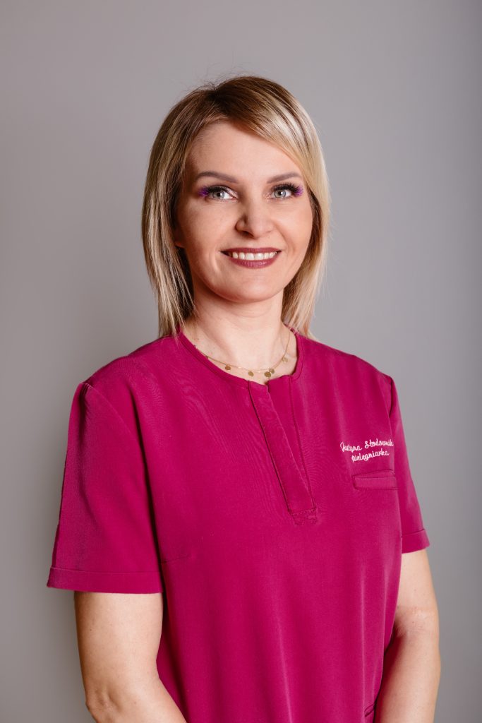 Pielęgniarka oddziałowa mgr Justyna Słodownik Specjalista pielęgniarstwa pediatrycznego. Kobieta w krótkich blond włosach, ubrana w różowy fartuch, na szarym tle.