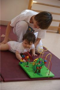 Zdjęcie przestawia leżące niemowlę, które bawi się drewnianą zabawką. Dziecko podtrzymuje mama. Znajdują się na sali gimnastycznej, w tle widać drabinki do ćwiczeń. 