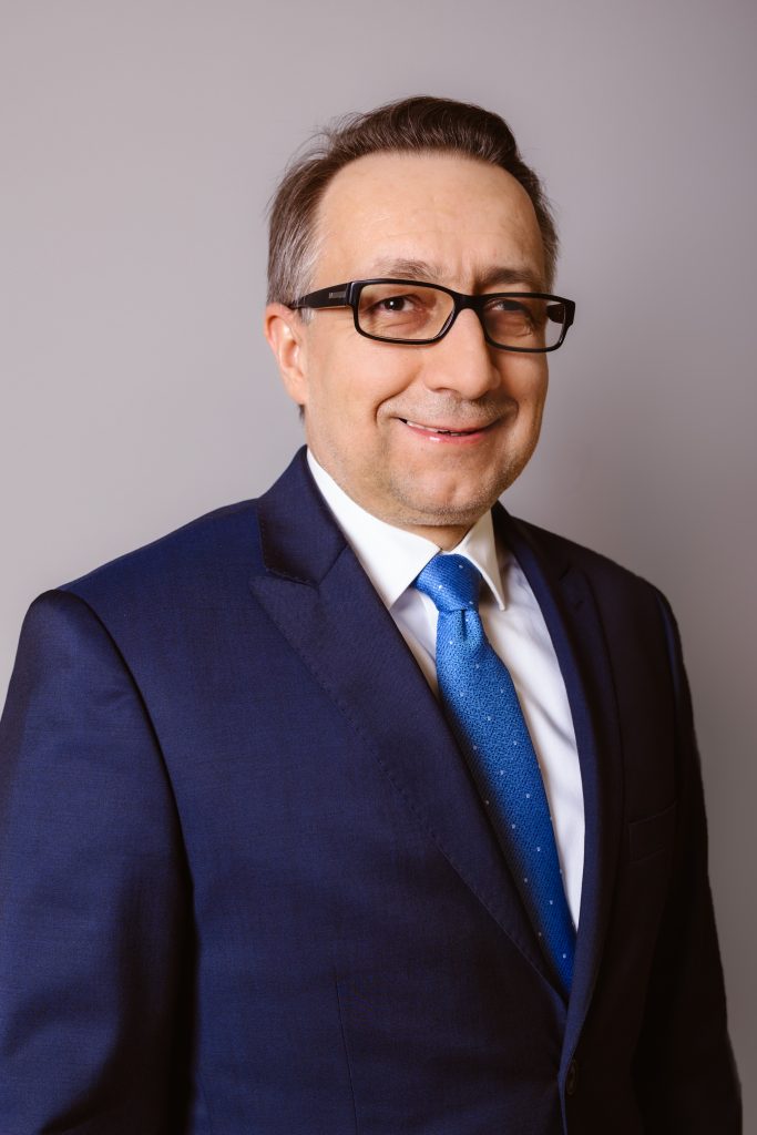 Dyrektor Szpitala dr inż. Roman Lewandowski. Mężczyzna w okularach, ubrany w ciemno niebieski garnitur, niebieski krawat, na szarym tle.
