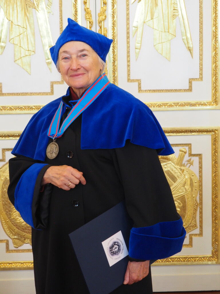 dr n.med. Krystyna Janukowicz. Kobieta ubrana w togę czarno-niebieską, stojąca na biało-złotym tle. na szyi zawieszony ma medal Gloria Medicinae 