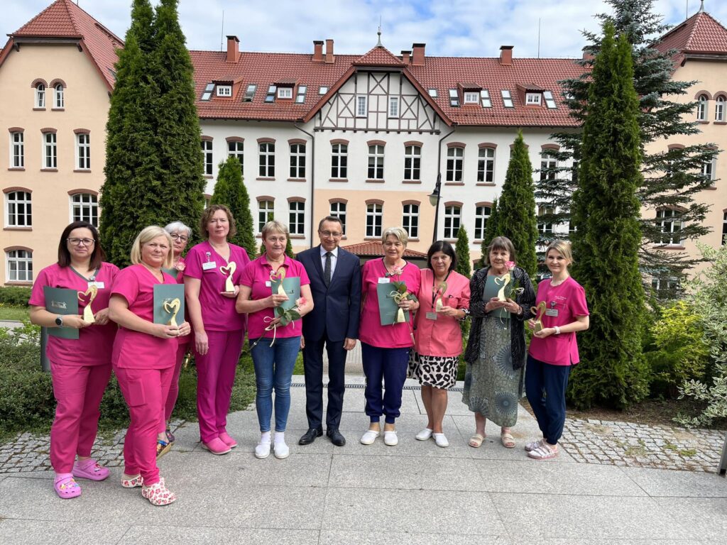 Dziewięć kobiet- pielęgniarek ubranych w różowe mundurki wraz z Dyrektorem Szpitala. Stoją przed budynkiem głównym szpitala. Pozują do zdjęcia,  widać zieloną roślinność przed szpitalem. 