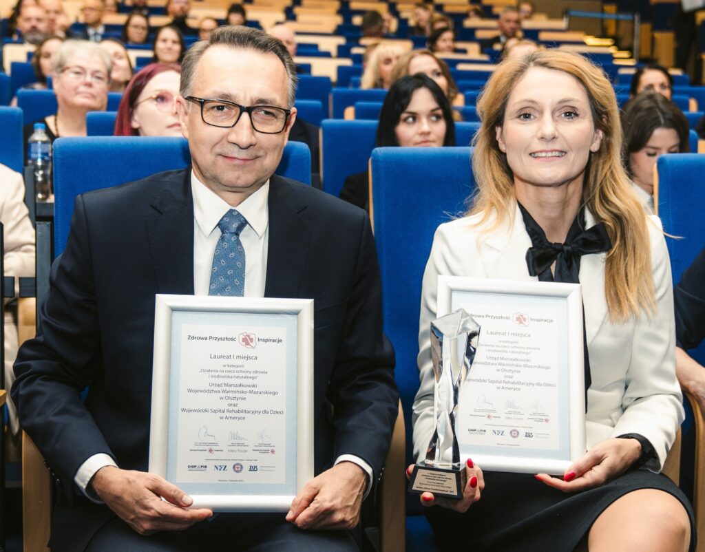 Na zdjęciu znajduje się dwóch uśmiechniętych laureatów nagród, siedzących w rzędzie w audytorium z niebieskimi fotelami. Mężczyzna po lewej stronie ma okulary, nosi ciemny garnitur i niebieski krawat, trzyma dyplom w białej ramce. Kobieta po prawej stronie ma długie blond włosy, jest ubrana w elegancką białą marynarkę z czarną kokardą pod szyją. Trzyma w dłoni statuetkę oraz podobny dyplom w białej ramce. W tle widać innych uczestników wydarzenia siedzących w kolejnych rzędach. Na dyplomach widoczne jest logo oraz napisy dotyczące nagrody „Zdrowa Przyszłość Inspiracje”, a także szczegóły dotyczące wyróżnienia w kategorii związanej z ochroną zdrowia.