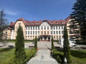 Na pierwszym planie schody i teren zielony szpitala, drzewa, krzewy, ławeczki. W tle budynek główny.