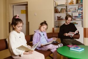 Trójka siedzących na ławce dzieci, znajdują się w pomieszczeniu. Dwie dziewczynki oraz chłopiec. Pierwsza dziewczynka ubrana jest w jasny dres, ma brązowe włosy, druga mniejsza dziewczynka ubrana jest w niebieską bluzkę, różową rozpinaną bluzę oraz różowe leginsy. Obok dziewczynek siedzi chłopiec ubrany w czarną bluzę i zielone spodnie. Każde dziecko czyta książę. Przed dziećmi stoi zielony stolik, na drugim planie widać ścianę na której znajdują się drzwi oraz szafa, która ma szklane drzwi. W szafie poukładane są książki oraz kartony z grami planszowymi.