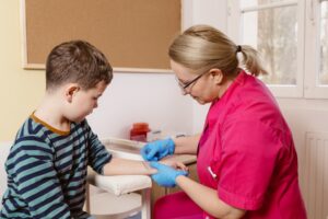 Pielęgniarka wykonująca badanie dla chłopca. Chłopiec w pozycji siedzącej ubrany w bluzkę w baski podaje rękę dla pielęgniarki. Pielęgniarka o blond włosach, ubrana w różowy fartuszek ma nałożone rękawiczki. Linijką dotyka rękę chłopca. W tle widać ścianę na której znajduje się korkowa tablica, przy ścianie stoi stolik na którym widać czerwony pojemnik.