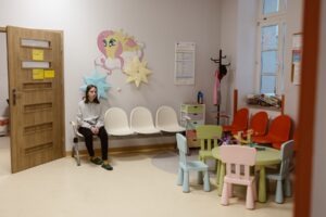 Poczekalnia poradni alergologicznej. W pomieszczeniu na ścianie namalowany jest biało różowy konik. Widać otwarte brązowe drzwi. Przy ścianach ustawione są krzesła, na krześle siedzi dziewczynka o ciemnych włosach, ubrana w białą bluzę i czarne spodnie. Po prawej stronie stoi mały stoliczek z kolorowymi krzesełkami. W rogu pomieszczenia ustawiony jest wieszak na ubrania oraz kolorowa szafka.