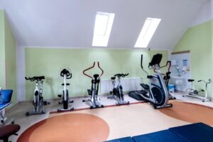 Sala gimnastyczna szpitala. Przy ścianie ustawionych jest 7 rowerów stacjonarnych. Po lewej stronie stoi niebieskie łóżko rehabilitacyjne, na podłodze rozłożone są niebieskie materace do ćwiczeń.