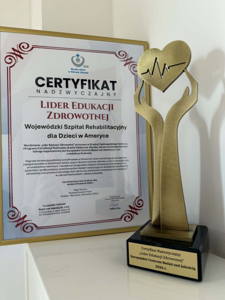 Na pierwszym planie stoi złota statuetka w kształcie rąk które trzymają serce, na dole statuetki napis Certyfikat Nadzwyczajny „Lider Edukacji Zdrowotnej”. Europejskie Centrum Badań nad Jakością 2026. W tle widać papierowy certyfikat włożony w złotą ramkę.